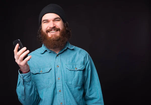 Happy bearded hipster man standing over dark background and holding smartphone — Stock Photo, Image