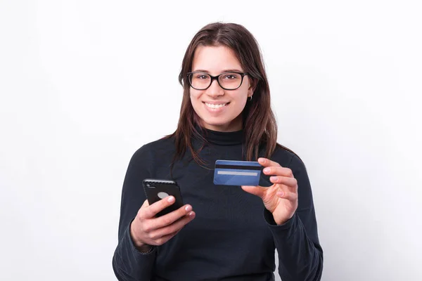 Neşeli genç bir kadın kameraya bakıyor ve elinde akıllı telefon ve kredi kartı tutuyor. — Stok fotoğraf