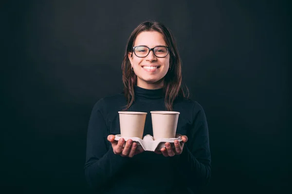 Jovencita alegre sobre fondo oscuro sosteniendo dos tazas de café para llevar — Foto de Stock