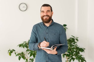 Smiling young bearded man in office making notes in planner