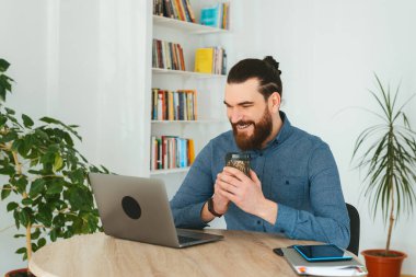 Neşeli sakallı adam elinde bir fincan kahve tutarken dizüstü bilgisayara bakıp gülümsüyor.