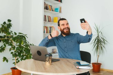 Ofisteki neşeli, yakışıklı, sakallı adam selfie çekiyor ve başparmağını gösteriyor.