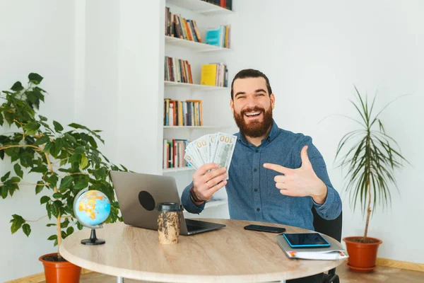 Cheerful young bearded office worker pointing at money — Stock Photo, Image