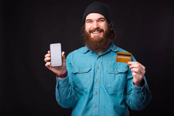 Retrato del hombre barbudo sonriente mostrando tarjeta de crédito y teléfono inteligente — Foto de Stock