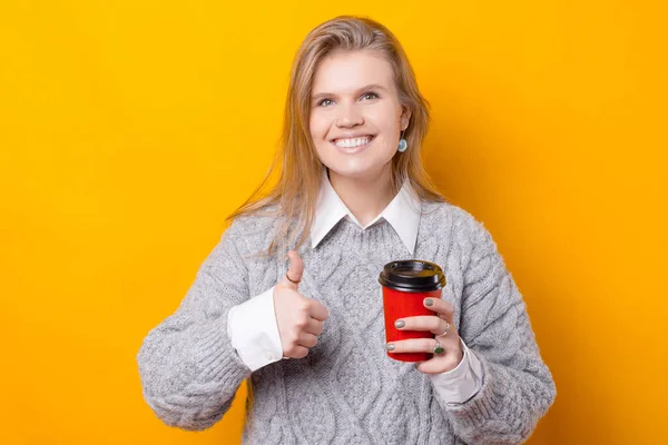 Молодая счастливая женщина держит чашку с горячим напитком и улыбается показывает большой палец вверх — стоковое фото