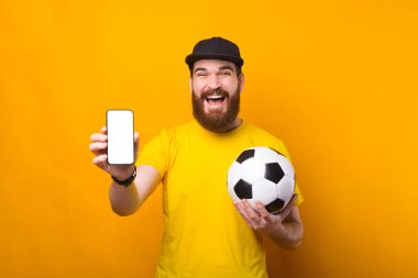 Heyecanlı genç bir adam elinde bir futbol topu ve gülümseyen bir telefon tutuyor.
