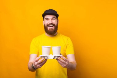 Neşeli genç sakallı adam sarı duvarın yanında iki bardak sıcak içecek tutuyor gülümsüyor