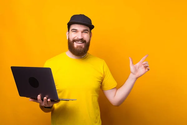 Barbudo joven feliz hombre está sosteniendo una computadora y que está apuntando a un espacio de copia — Foto de Stock