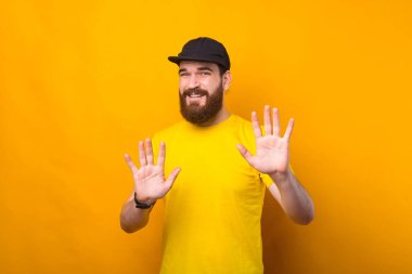 Sakallı genç bir adam sarı bir duvarın yanında duruyor. Dur işaretini gösteriyor.