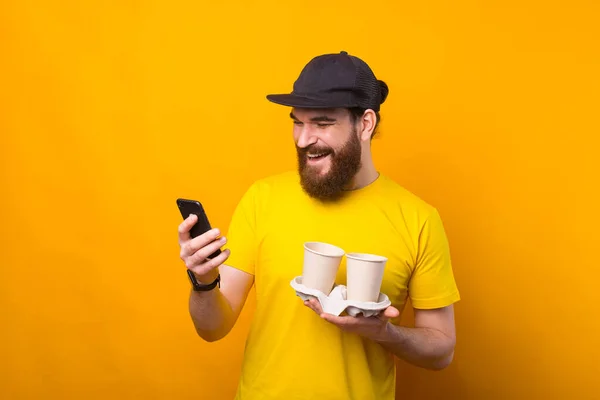 Foto de un alegre hombre hipster barbudo con camiseta amarilla sosteniendo dos tazas de café para llevar y usando el teléfono — Foto de Stock