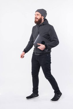 Beyaz arka planda elinde dizüstü bilgisayarla dolaşan sakallı bir adamın tam boy fotoğrafı.