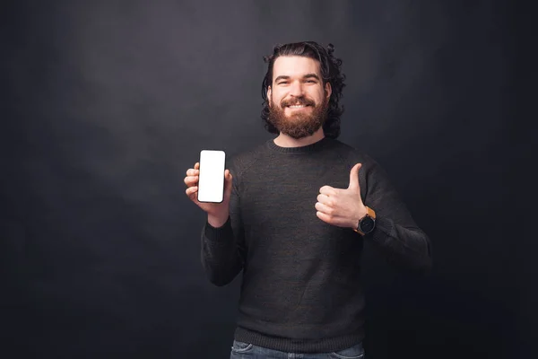 Foto de hombre barbudo guapo mostrando la pantalla en blanco en el teléfono móvil y mostrando el pulgar hacia arriba — Foto de Stock