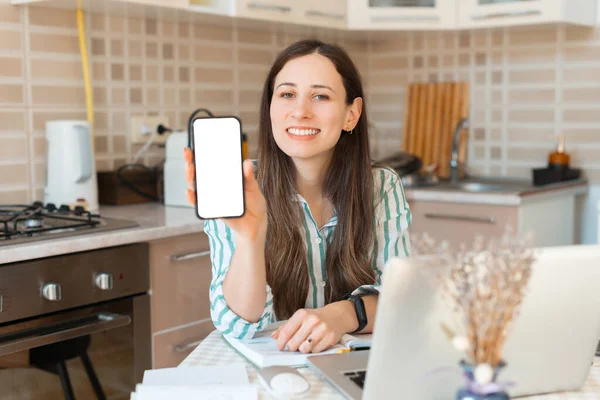 Foto de una joven feliz trabajando desde casa y mostrando la pantalla en blanco del teléfono inteligente — Foto de Stock