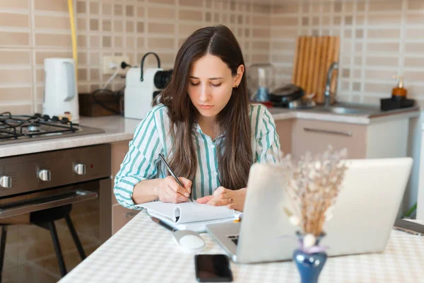 Photo de jeune femme prenant des notes dans le planificateur assis sur la table dans la cuisine — Photo