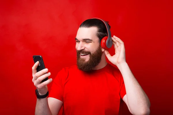 Joven alegre hombre barbudo feliz escuchando música sobre fondo rojo —  Fotos de Stock