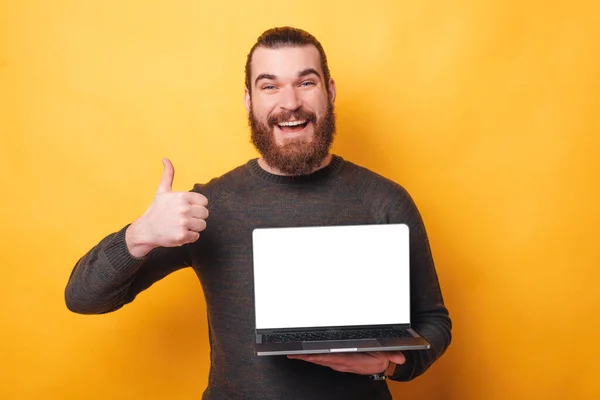 Beau jeune homme avec barbe montrant pouce vers le haut et montrant écran vierge sur ordinateur portable — Photo