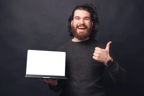 Веселий красивий чоловік з бородою в повсякденному житті показує великий палець вгору і ноутбук з порожнім екраном копія простору — стокове фото
