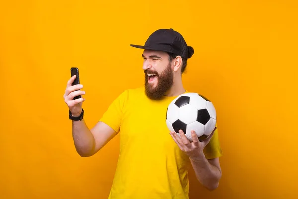 Портрет здивованого бородатого юнака, який дивиться на смартфон і тримає футбольний м'яч, підтримує улюблену команду — стокове фото