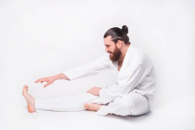 Tekvando üniforması giyen sakallı bir adamın beyaz arka planda esnediği bir fotoğraf.