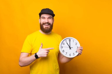 Yakışıklı, mutlu, gülümseyen sakallı hippi koca beyaz saati işaret ediyor.