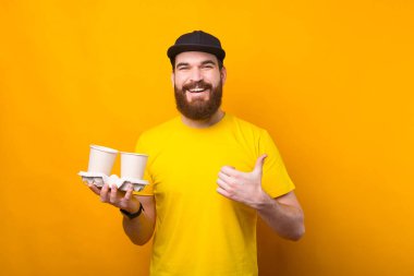 Çekici sakallı adam baş parmağını gösteriyor ve sarı arka planda kağıt kahve bardakları tutuyor.