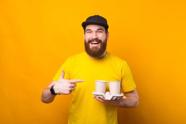 Mutlu sakallı hippi, sarı arka plandan uzaklaşmak için iki kahve bardağını işaret ediyor.