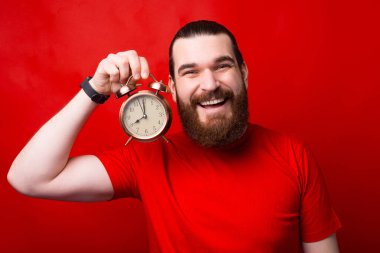 Photo of cheerful young bearded man holding alarm clock over red background.
