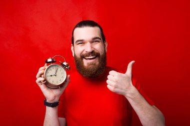 Photo of smiling young bearded man showing thumb up and alarm clock.