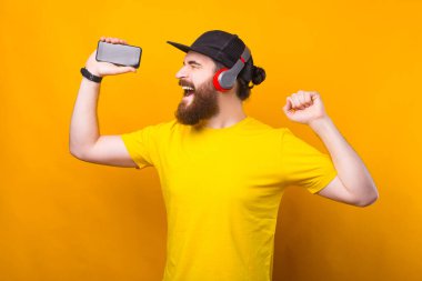 Sakallı neşeli hipster adamın kablosuz kulaklıklarda müzik dinlediği ve elinde akıllı telefon tuttuğu fotoğraf..