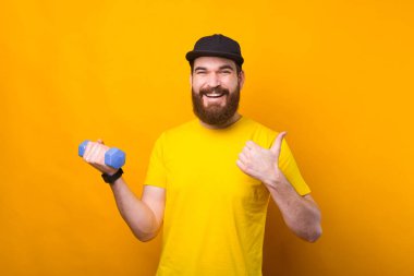 Mutlu gülümseyen sakallı genç adam başparmağını gösteriyor ve küçük bir halterle çalışıyor..