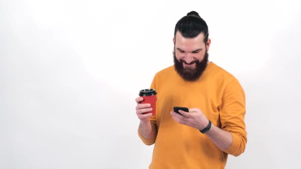 Beelden van de jonge man met de baard drinken van een kopje koffie te gaan en het gebruik van smartphone — Stockvideo