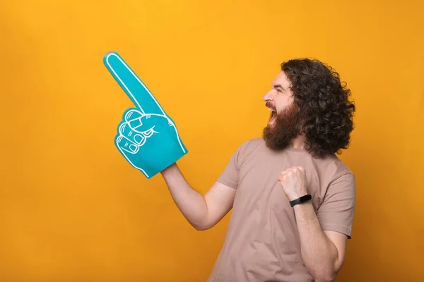 Amazed young bearded man pointing away with fan foam glove over yellow background.
