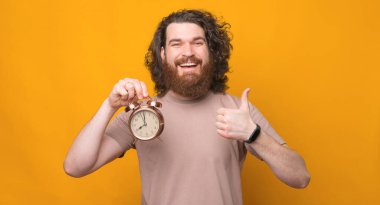 Uzun saçlı, neşeli, sakallı bir gencin portresi baş parmak hareketi ve çalar saat gösteriyor..