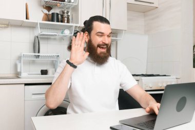 Neşeli genç sakallı hippi adam mutfakta oturuyor ve web kamerasında konuşuyor..