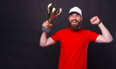 Şaşırmış genç sakallı adam zaferi kutluyor ve siyah arka planda şampiyonluk kupasını tutuyor..