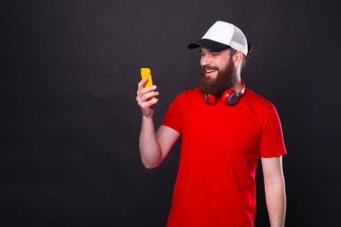 Siyah arka plan yakınlarında sakallı, gülümseyen genç bir adamın fotoğrafı. Akıllı telefon kullanıyor..