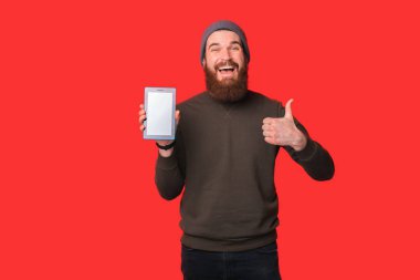 Şaşırmış sakallı hippi adam kırmızı arka planda baş parmağını ve tabletini gösteriyor..