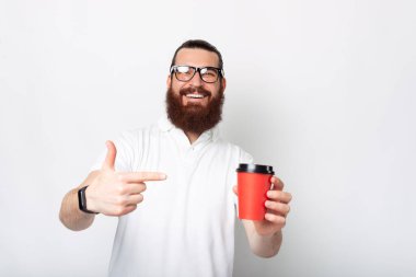 Olumlu gülümseyen sakallı adam beyaz arka planda bir bardağı işaret ediyor..