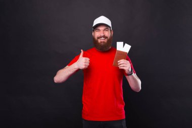 Genç sakallı adam pasaportunu ve iki seyahat biletini elinde tutarken siyah arka planda baş parmağını gösteriyor. İyi organize edilmiş bir gezi..