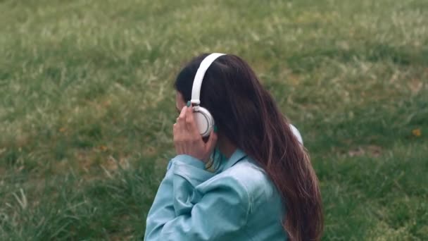 Beelden van jonge zakenvrouw zittend op gras in park en luisterend naar muziek — Stockvideo