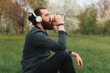 Genç bir adamın kulaklıkla doğanın tadını çıkarırken ve çimenlerde oturup kahve içerken çekilmiş güzel bir fotoğrafı. .