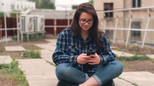 Bilder på ung kvinna som sitter på taket och använder smartphone över staden bakgrund — Stockvideo