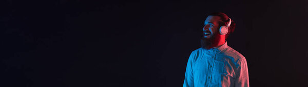 Фото улыбающегося бородатого хипстера на темном фоне с синим и красным неоновым светом, слушающего музыку в наушниках.