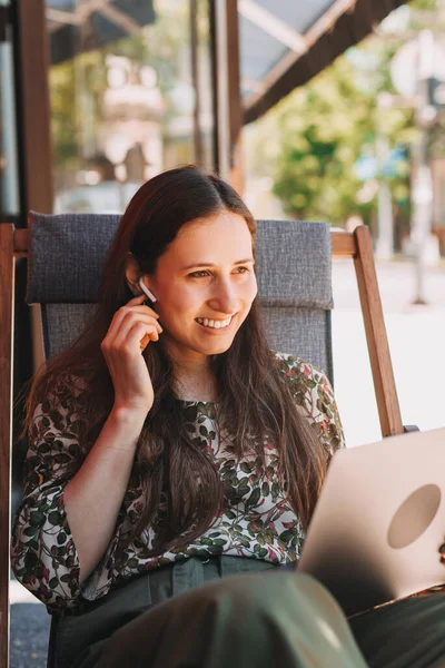 Retrato de una joven sentada al aire libre en la cafetería y usando auriculares y portátil —  Fotos de Stock