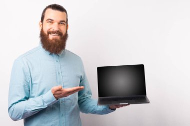 Sıradan mavi gömlekli yakışıklı bir adam dizüstü bilgisayarında boş ekranı gösteriyor ve beyaz duvardan gülümsüyor..