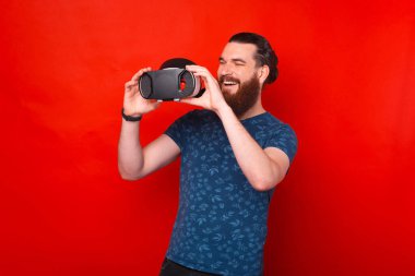 Kızıl zemin üzerinde sanal gerçeklik kaskı tutan sakallı genç adamın fotoğrafı..