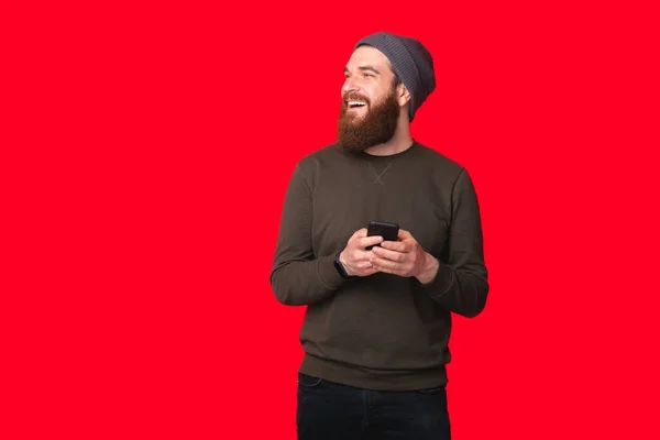 Uśmiechnięty Brodaty Hipster Używa Smartfona Odwraca Wzrok Czerwonym Tle — Zdjęcie stockowe