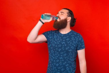 Kırmızı arka planda duran şişeden su içen sakallı genç adamın portresi..