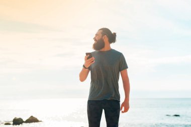 Elinde cep telefonu olan yakışıklı sakallı adam, mesaj atıyor ve tropikal arka plana bakıyor..
