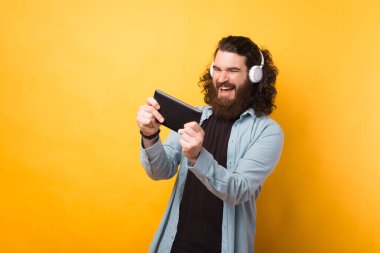 Hefon takan ve sarı arka planda akıllı telefondan oynayan heyecanlı sakallı adamın fotoğrafı.